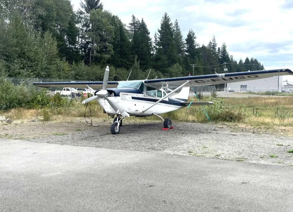 A white and blue Cessna 185 aircraft with registration C-FUGG parked on a gravel patch next to an asphalt path, with trees in the background.