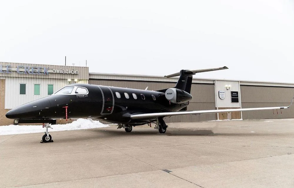 A black Embraer Phenom 300 jet (N506P) parked on a snowy tarmac in front of an Eagle Creek Aviation hangar.