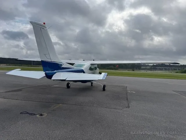 The rear view of a white and blue Cessna 182 is shown parked on an asphalt tarmac under a cloudy sky.