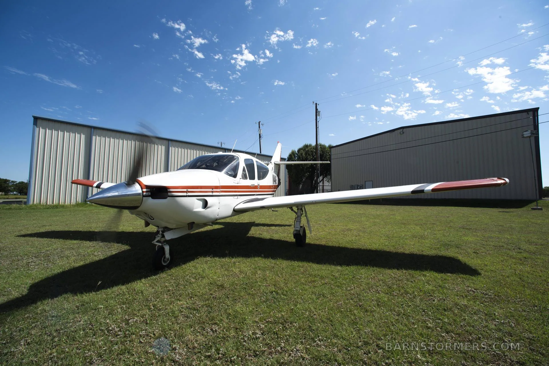 A white and red single-engine aircraft with a spinning propeller sits on a grassy field in front of hangars under a blue sky.