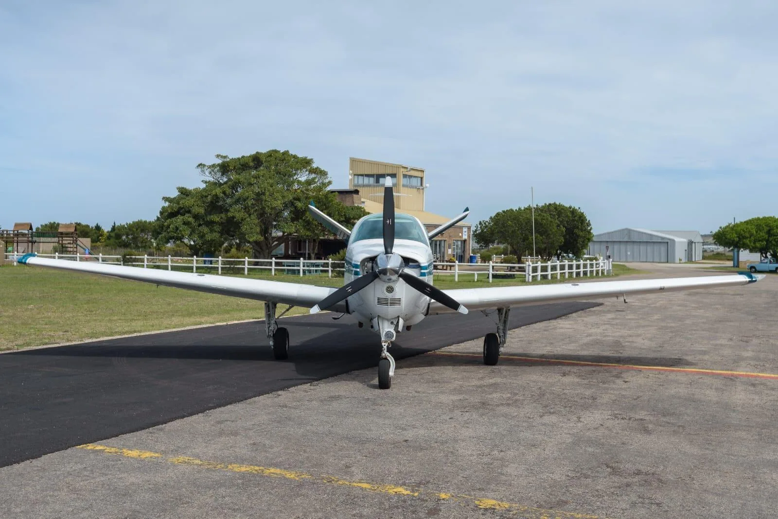 A white single-engine propeller aircraft with blue stripes is parked on an asphalt tarmac at an airfield, viewed from the front.