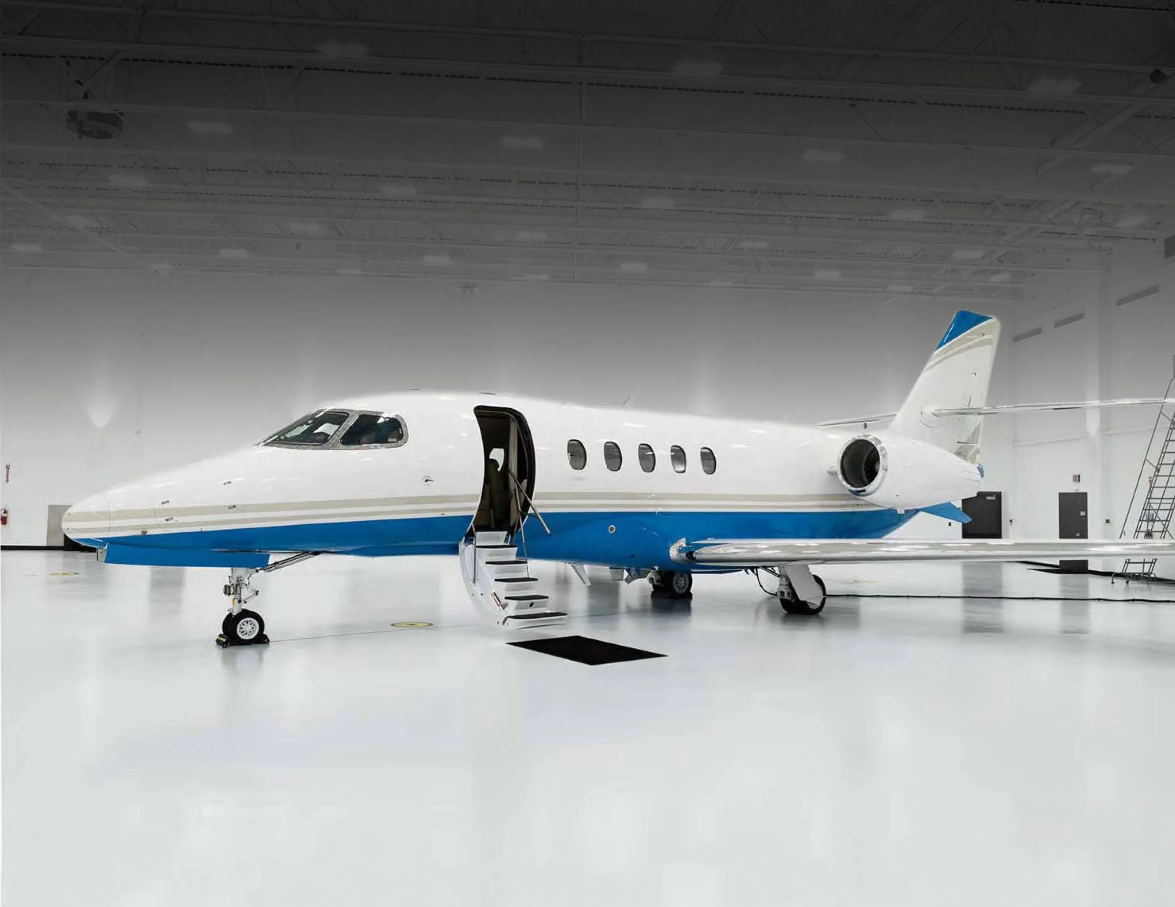 A white and blue business jet with its door open and stairs extended, parked inside a brightly lit hangar.