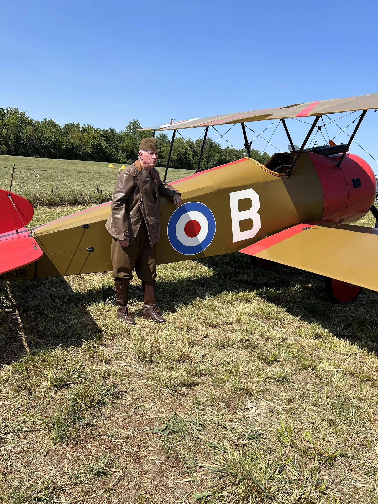 A man in a brown leather flight suit stands next to a tan and red biplane with British roundels on a grassy field.
