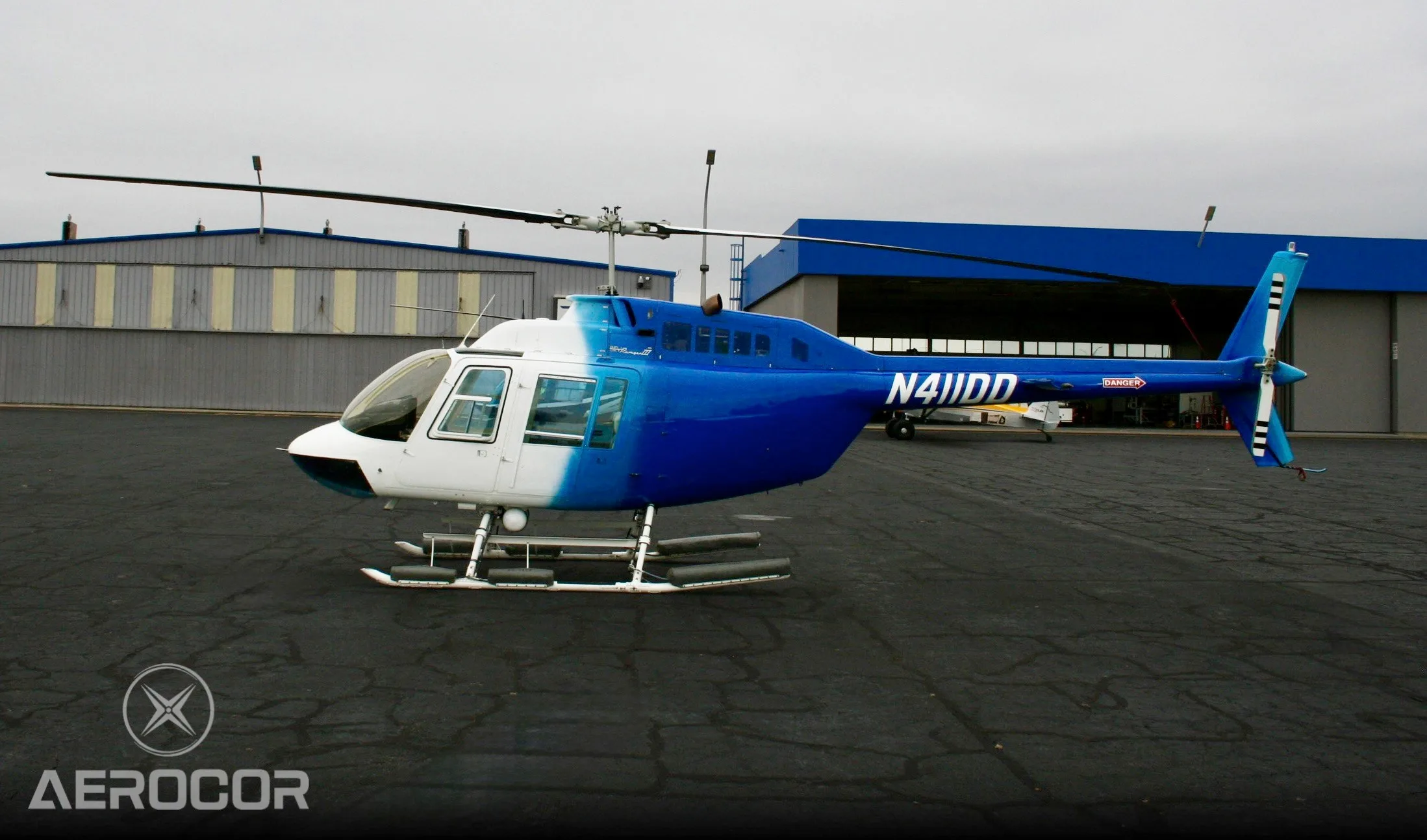 A blue and white Bell JetRanger III helicopter, tail number N411DD, parked on an airport tarmac with hangars in the background.