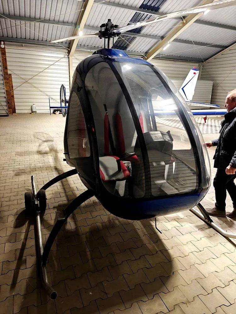 The front view of a blue single-seat helicopter with a clear canopy and red and gray seats, parked inside a hangar next to a person.