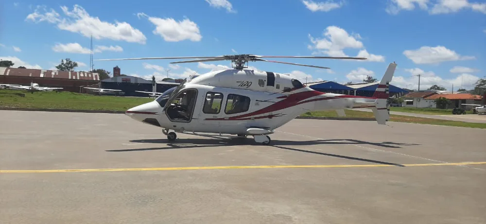 A white Bell 429 helicopter with red and grey stripes parked on an asphalt tarmac under a blue sky.