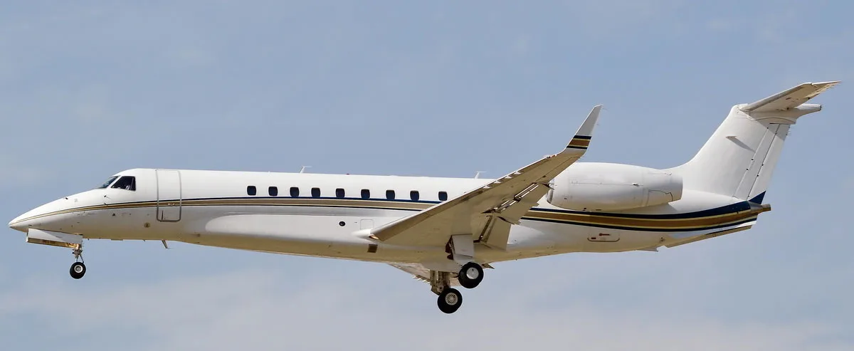 A white Embraer Legacy business jet with gold and blue stripes on its fuselage and tail, flying with landing gear extended against a light blue sky.