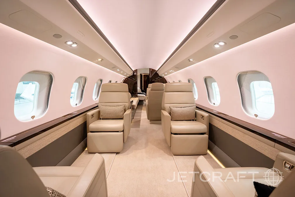 The luxurious interior cabin of a private jet features light beige leather seats, a cream carpet, and soft ambient lighting.