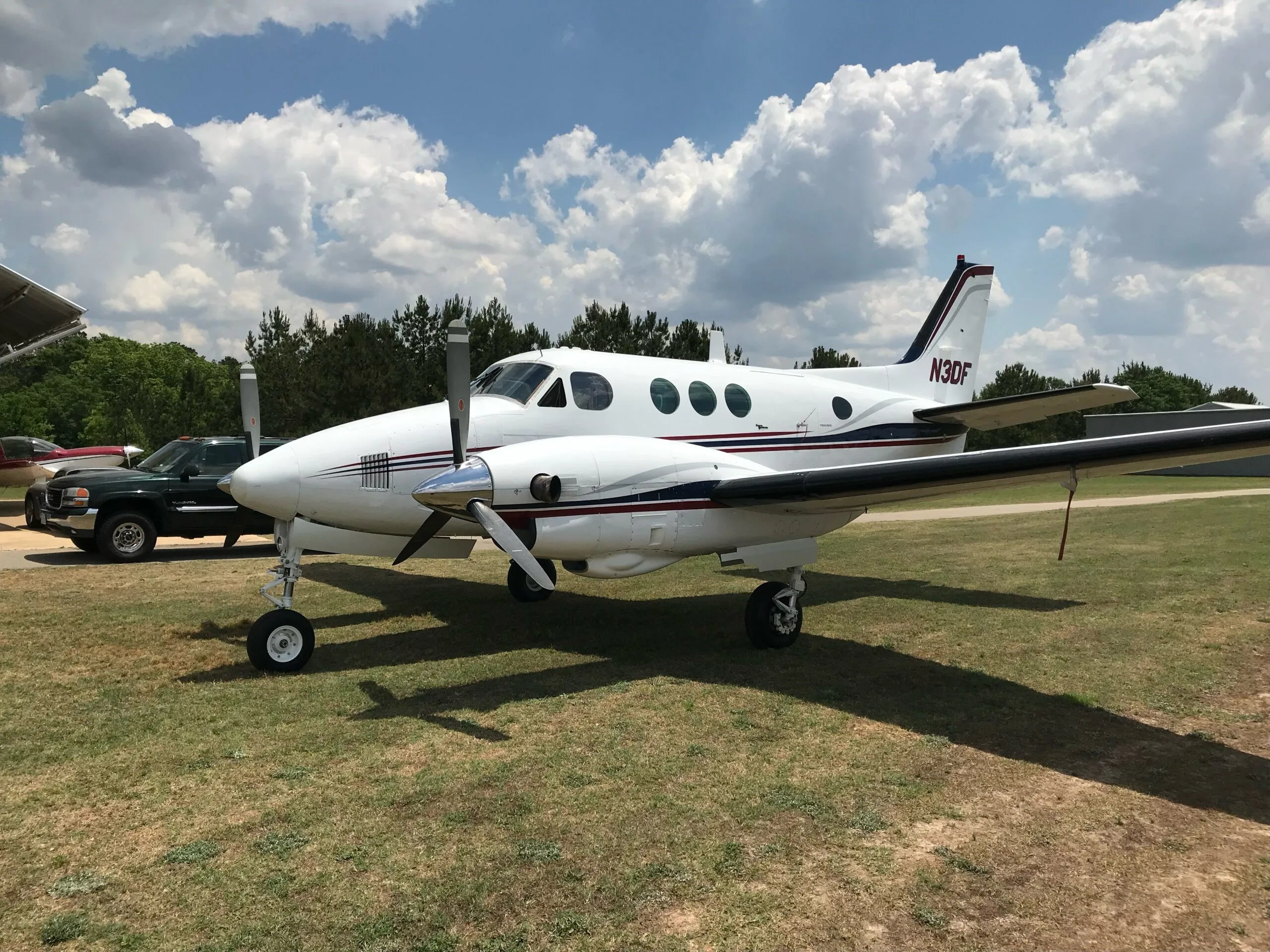 A white twin-engine turboprop aircraft, N3DF, with red and blue stripes, parked on a grassy field under a partly cloudy sky.