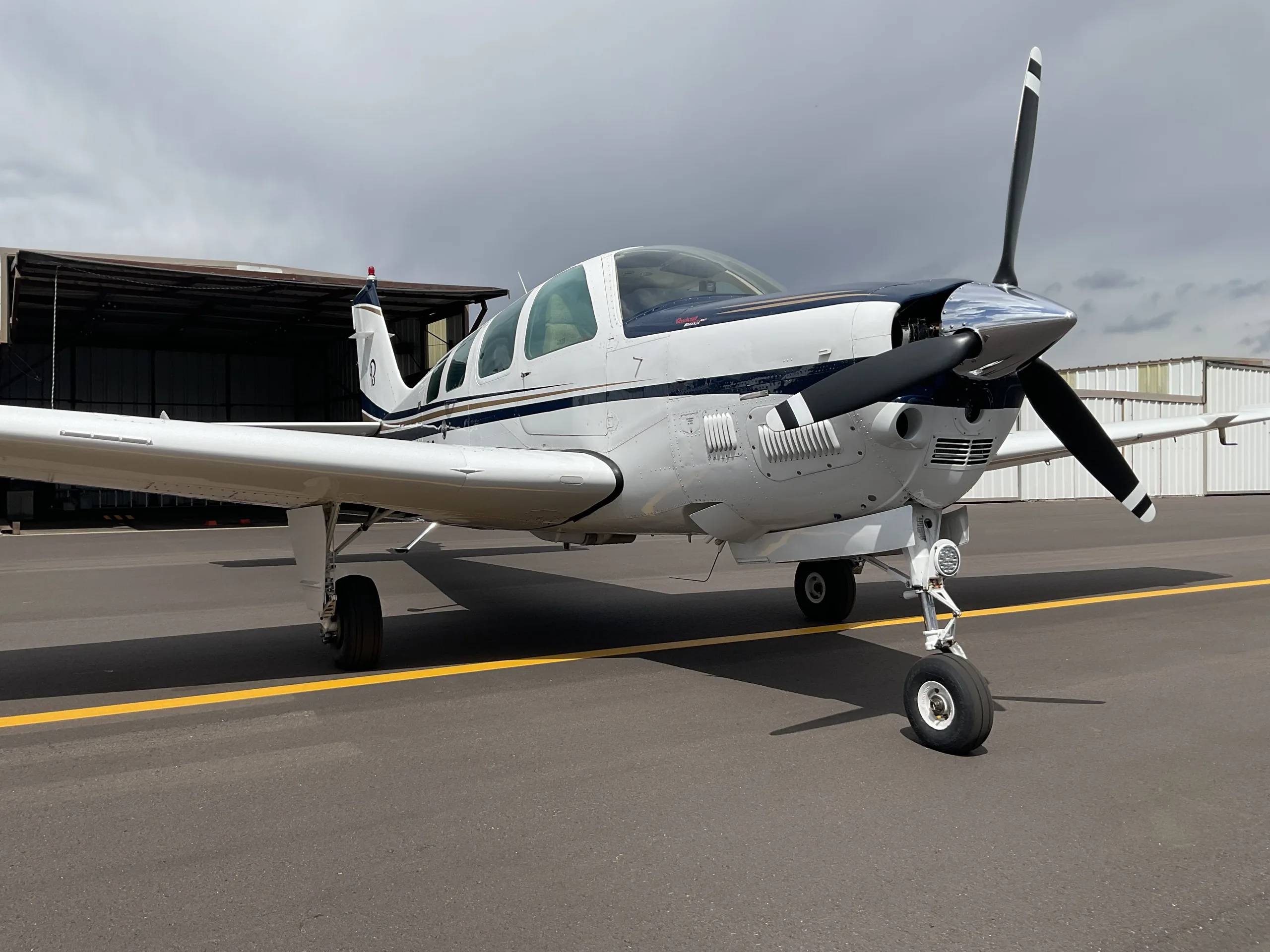 A white and blue Beechcraft Bonanza with a V-tail is parked on an asphalt tarmac under a cloudy sky.