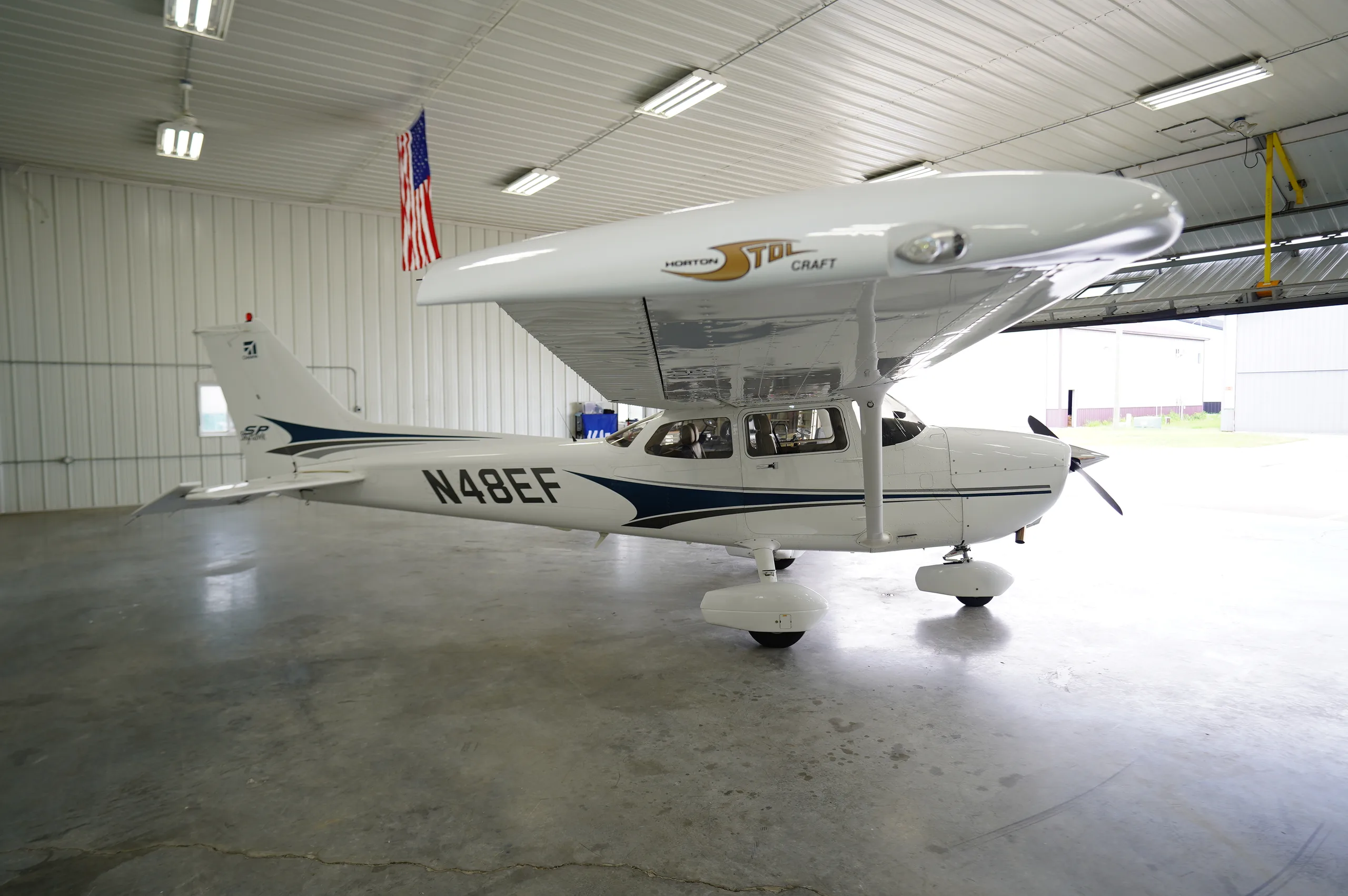 Side view of a white Cessna 172 (N48EF) with blue and black stripes in a hangar with the door open.
