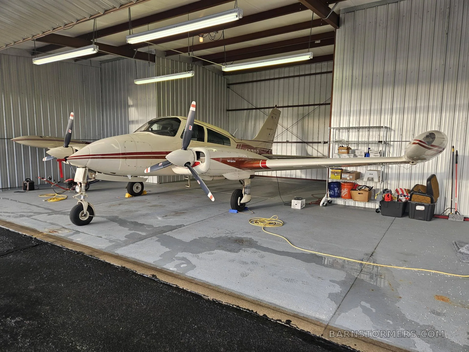 A white and red twin-engine propeller aircraft is parked inside a hangar, viewed from the front-right side.