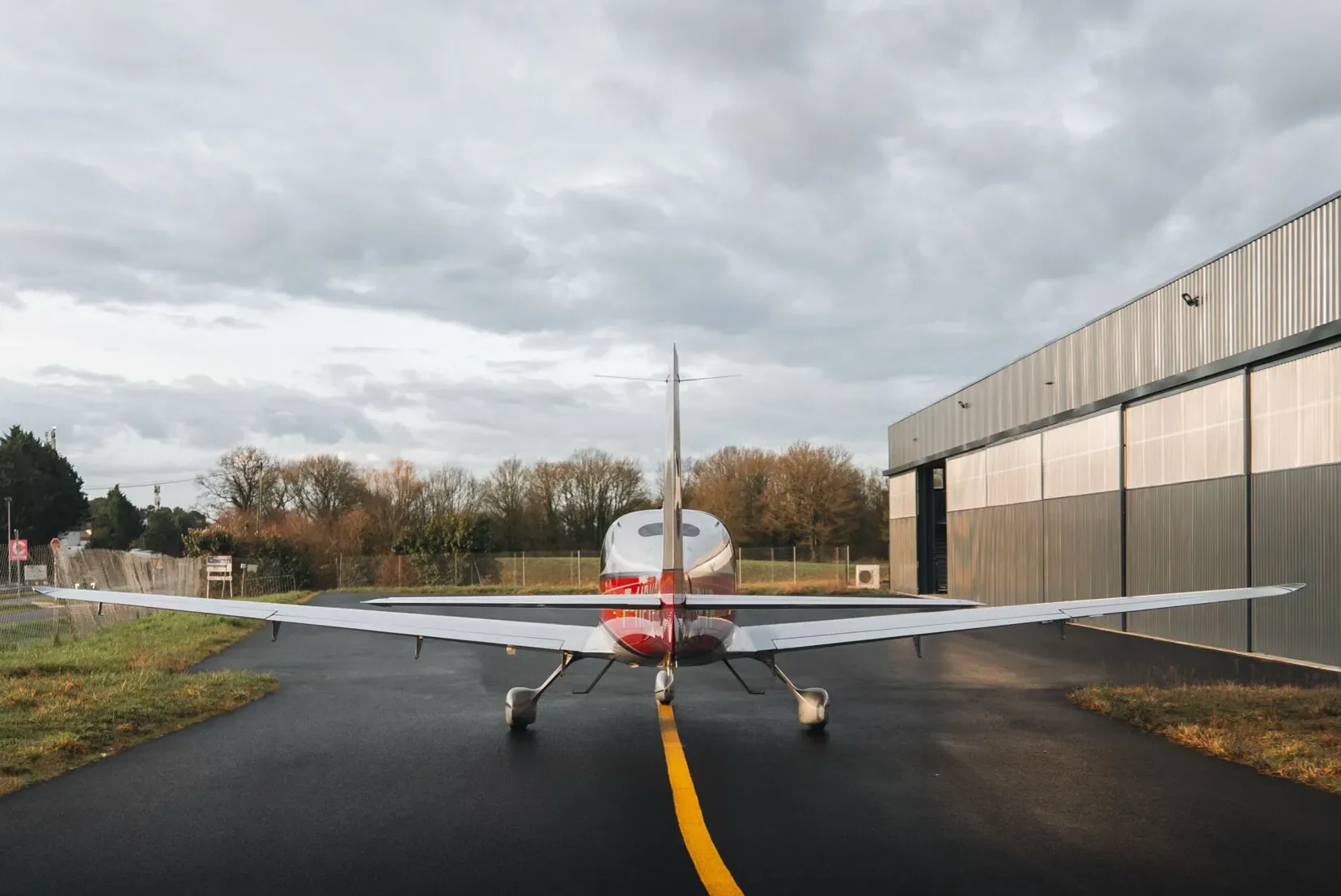 A silver and red Cirrus SR22 aircraft viewed from the rear, parked on an airfield next to a hangar.