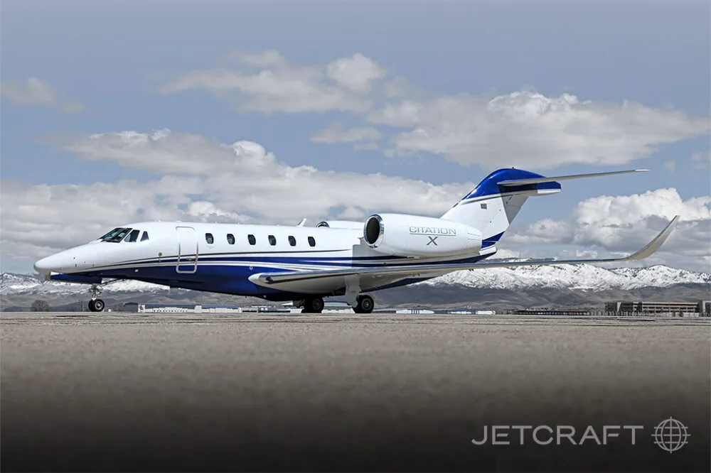 A white and blue Cessna Citation X business jet is parked on an airfield with snow-capped mountains in the background.