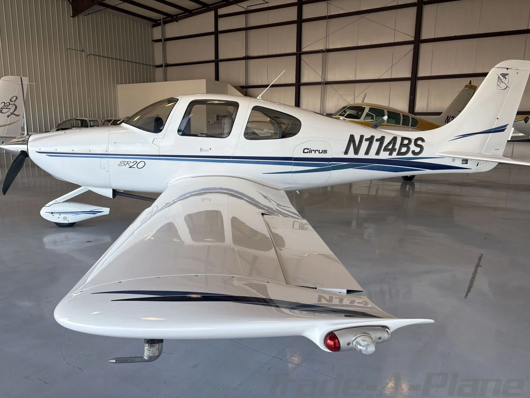 A white Cirrus SR20 aircraft with blue stripes, registration N114BS, parked in a hangar.