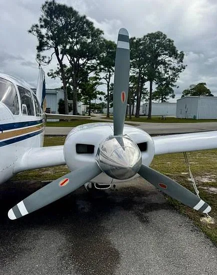A close-up view of the shiny propeller spinner and gray blades of a twin-engine aircraft, with trees and hangars in the background.