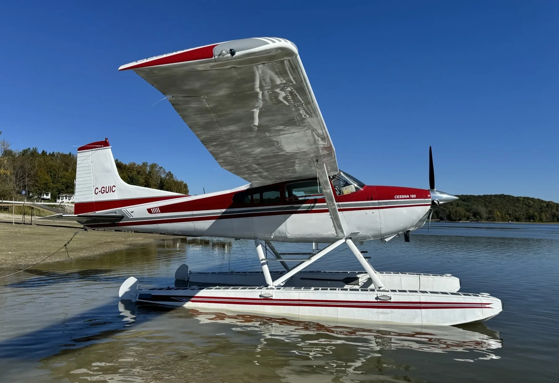 A red and white Cessna 185 floatplane is seen from the side, partially in water near a tree-lined shore.