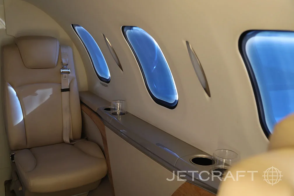 The private jet cabin shows beige leather seats, multiple windows, and a side console with cup holders.