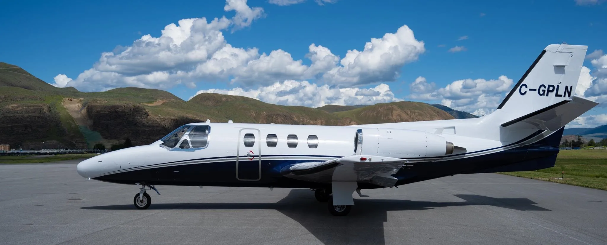 A white and dark blue business jet, registration C-GPLN, is parked on an airfield with mountains and a blue sky with clouds in the background.