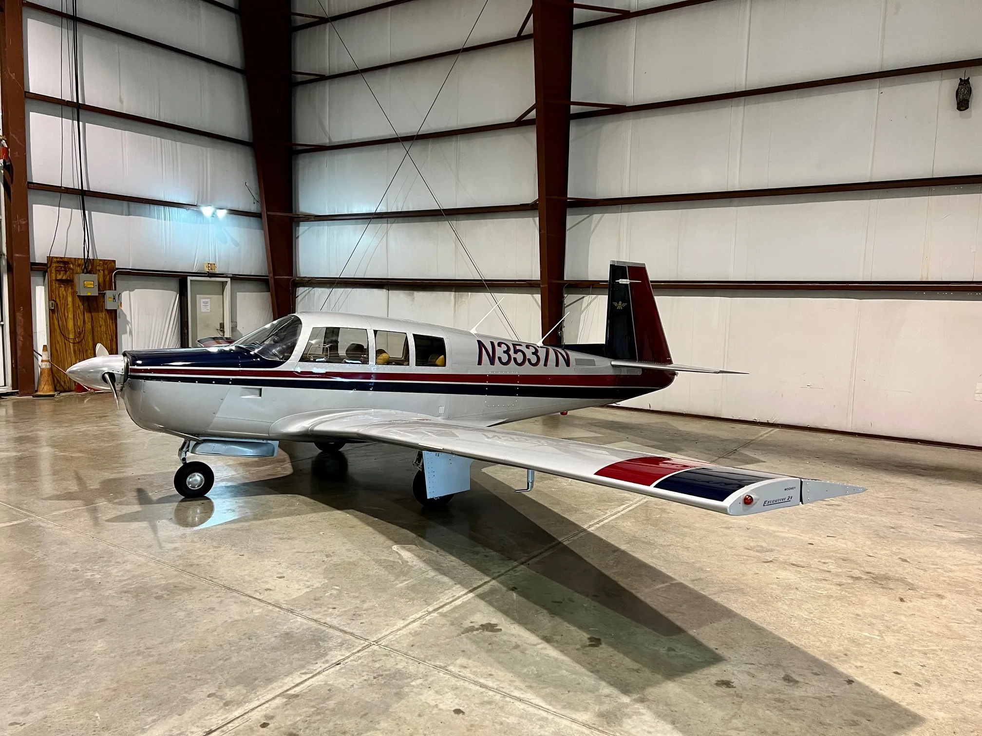 A silver single-engine Mooney Executive 21 aircraft with blue and red stripes, parked inside a hangar.