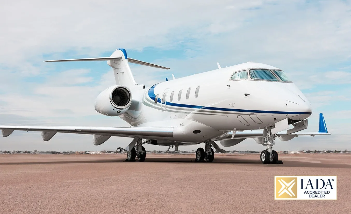 A white business jet with blue and grey stripes is parked on an airfield under a partly cloudy sky.