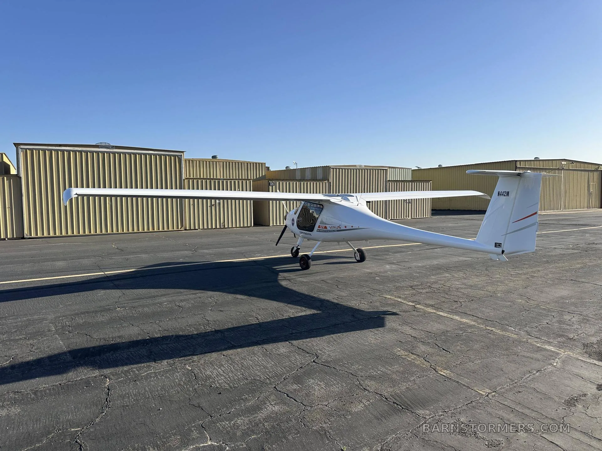 A white light sport aircraft, N442M, with orange stripes on the tail, viewed from the rear-left on an asphalt tarmac.