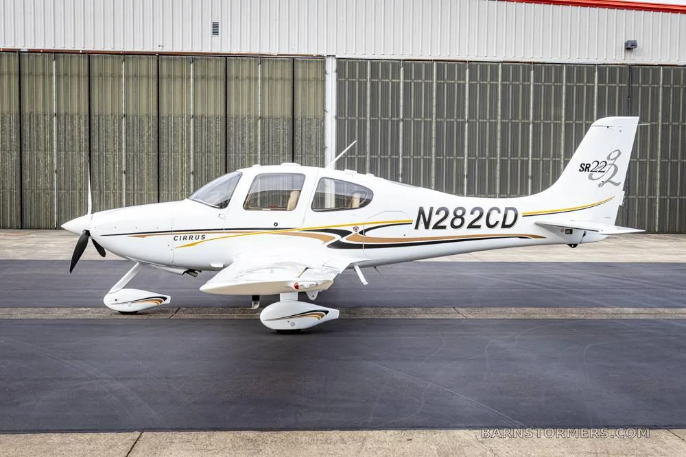 A white Cirrus SR22 aircraft with black and gold stripes is parked on an asphalt tarmac in front of a hangar.