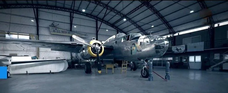 A wide shot of a silver B-25 Mitchell bomber with yellow propeller hubs, parked in a large hangar.