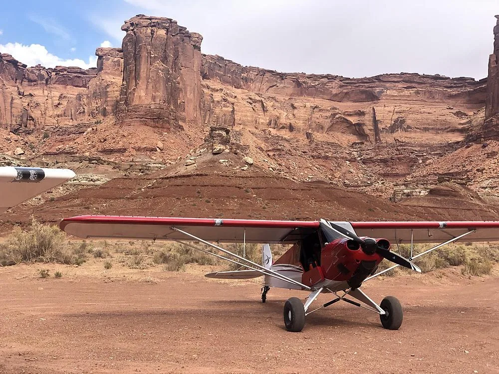 A red and white bush plane with large tires parked on a dirt strip in a desert landscape with large rock formations.