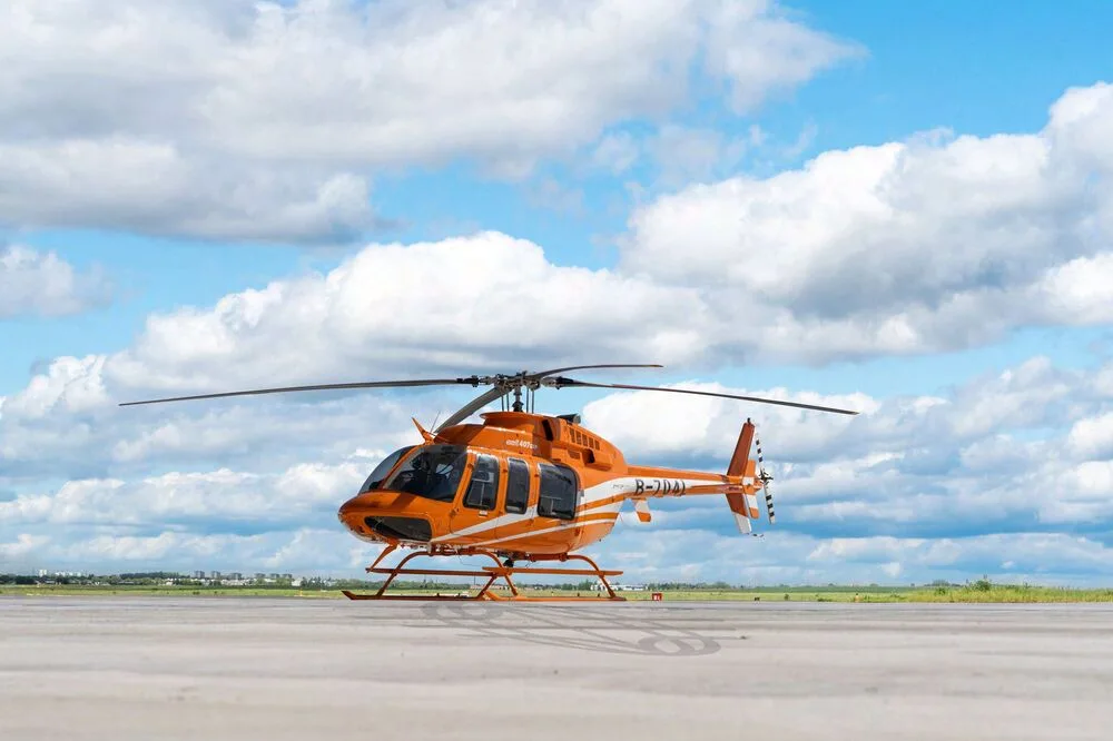 A bright orange Bell 407EXP helicopter with registration B-704L sits on an airfield under a cloudy sky.