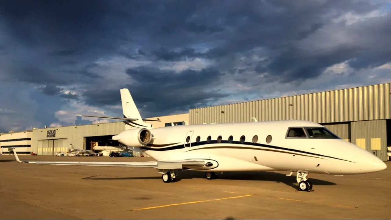 A white business jet with dark and gold stripes parked on an airfield in front of hangars under a dramatic cloudy sky.