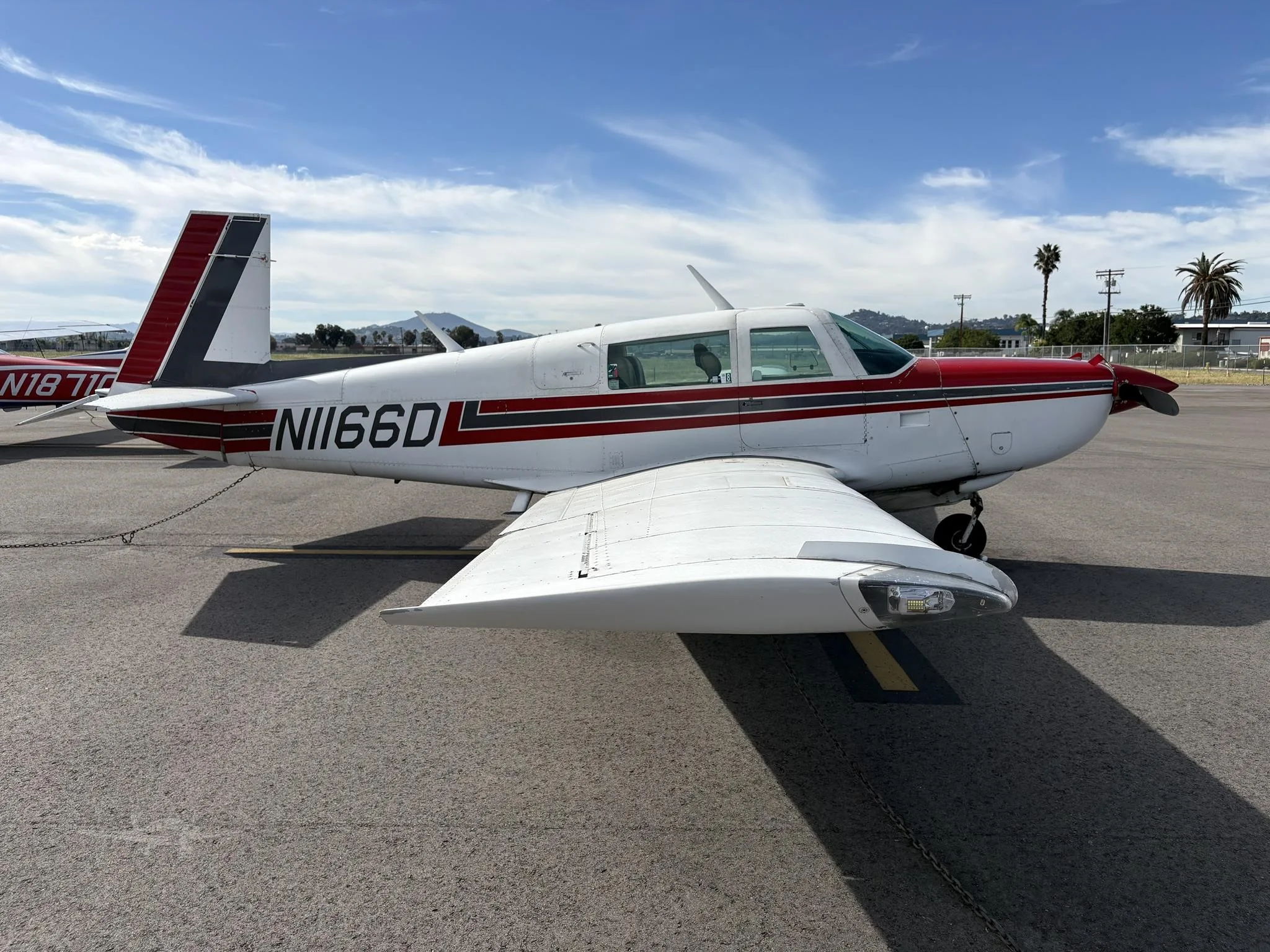 A white single-engine propeller aircraft with red and black stripes is parked on an airfield under a blue sky.