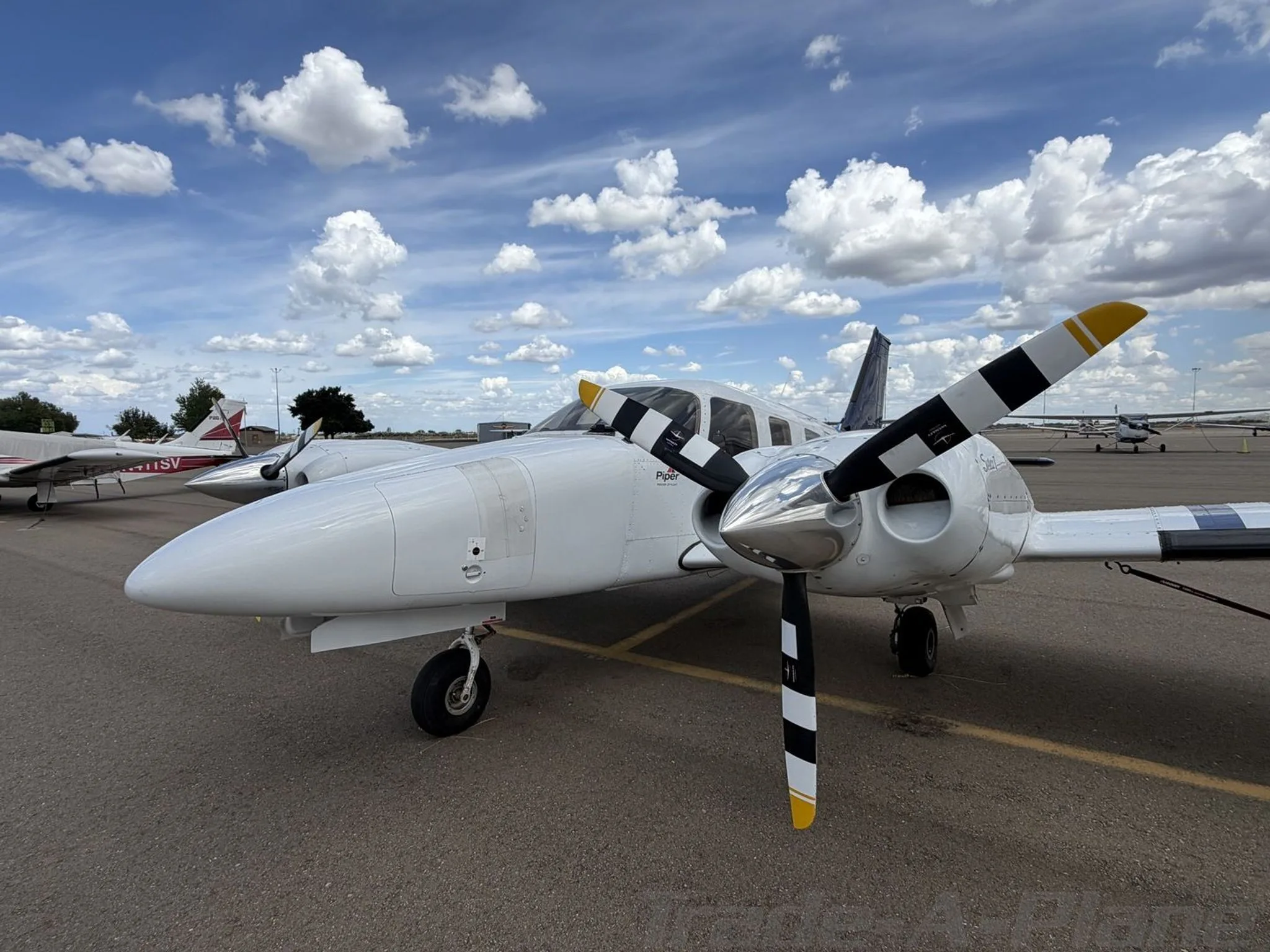 A low-angle front-right view of a white Piper Seneca II twin-engine aircraft with black and yellow striped propellers on an airport tarmac under a partly cloudy sky.
