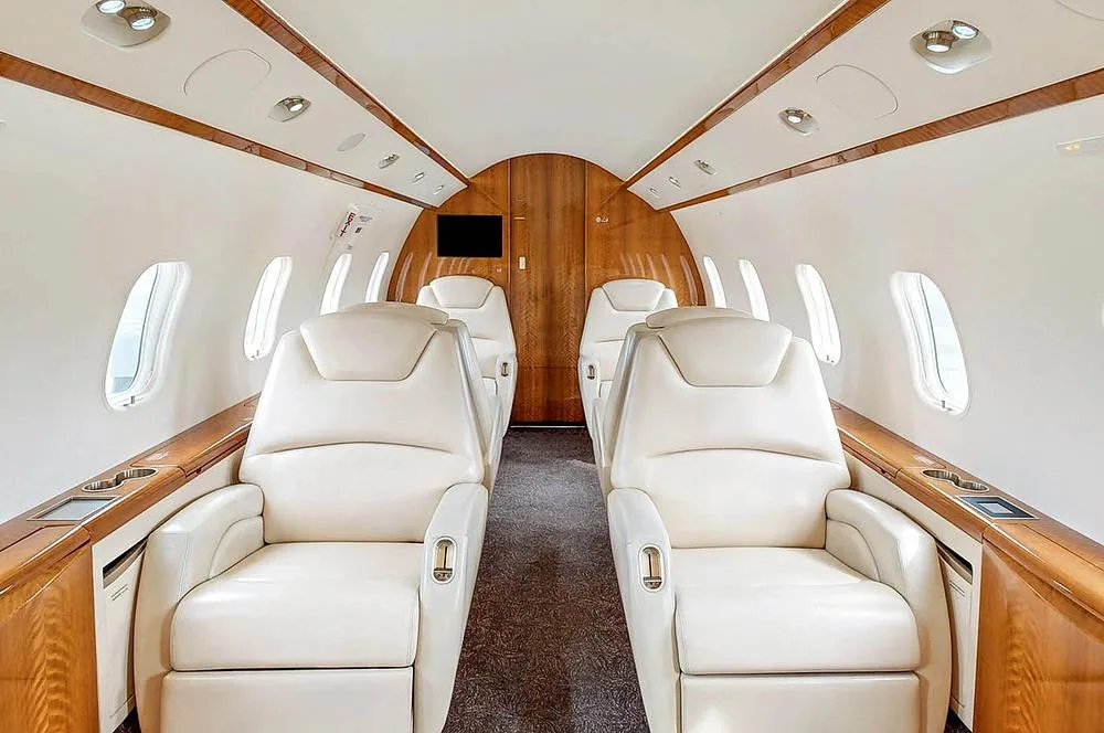 The aircraft cabin interior with light cream leather seats, wood paneling, and a dark carpet.