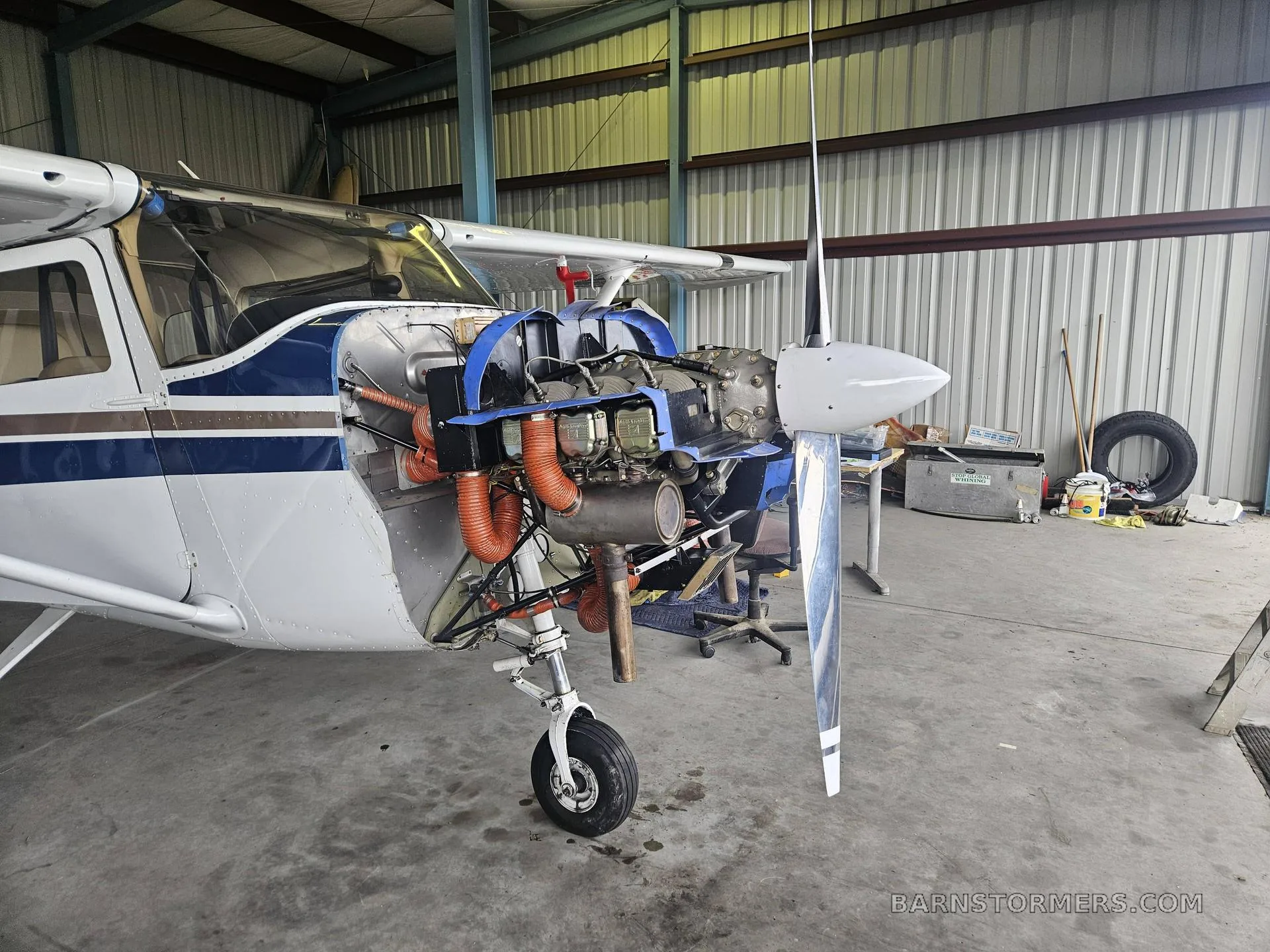A white and blue single-engine aircraft in a hangar with its engine cowling removed, revealing the engine components.