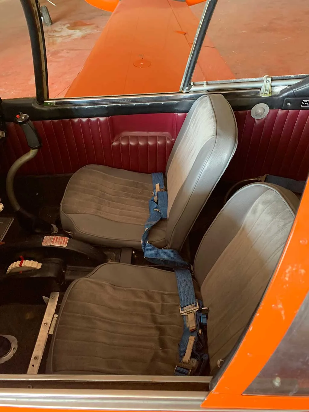 The aircraft's interior features two grey bucket seats with blue seatbelts and red side panels.
