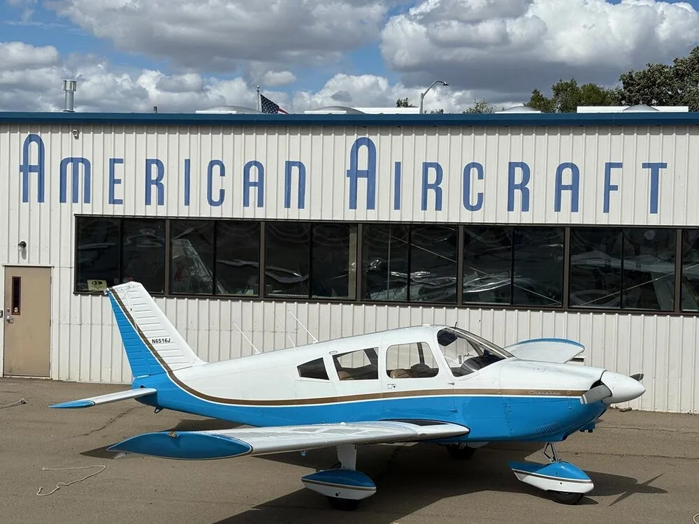 A white and blue Piper Cherokee D aircraft (N6516J) is parked in front of an "AMERICAN AIRCRAFT" hangar.
