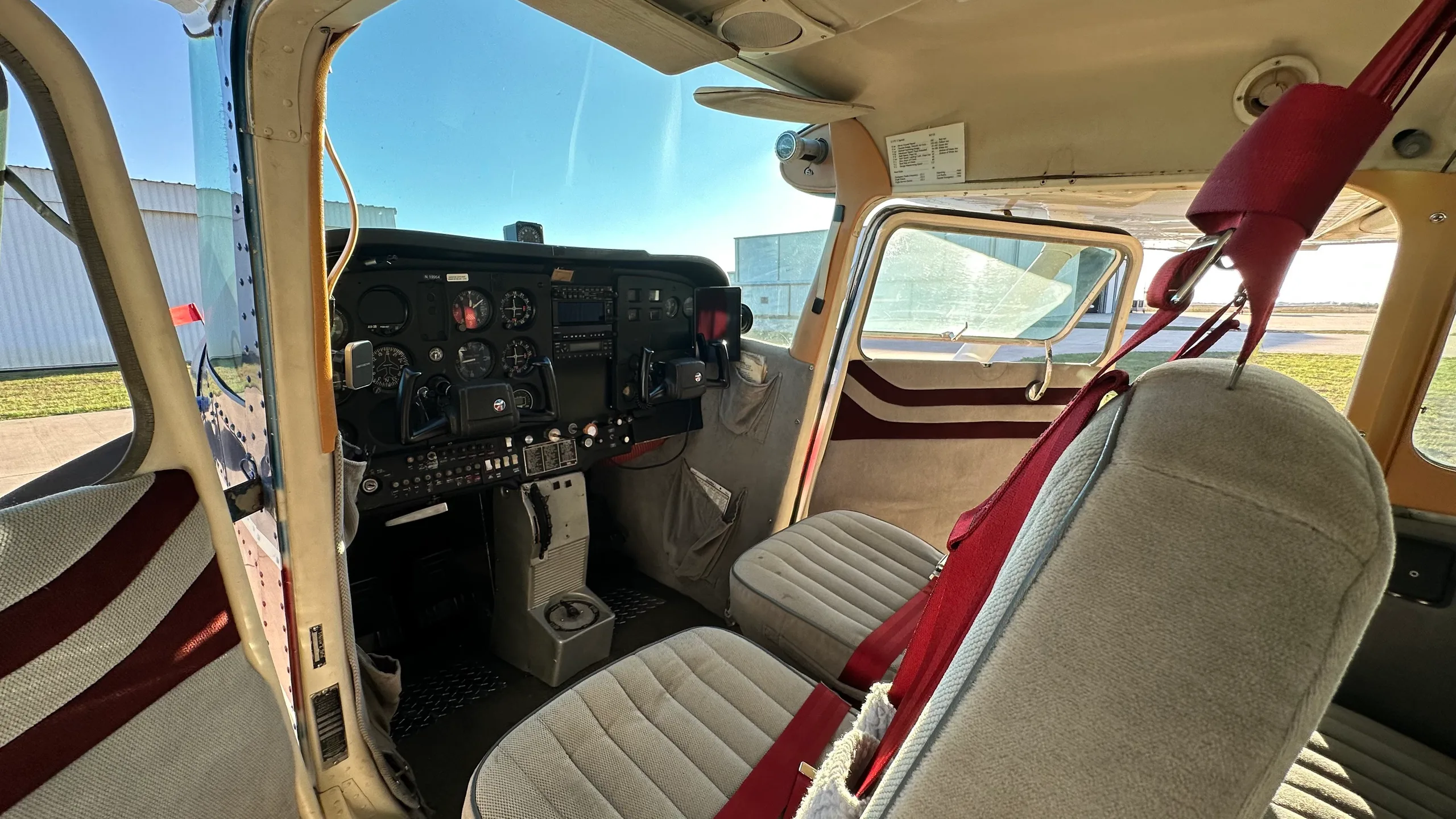 The interior of a small aircraft cabin with two front seats featuring red seatbelts and a view of the cockpit and outside.