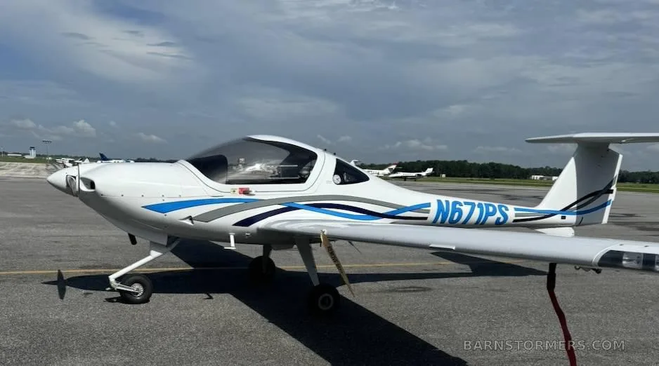 A white Diamond DA20-C1 Eclipse aircraft with blue and grey stripes, registration N671PS, parked on an airport tarmac under a cloudy sky.