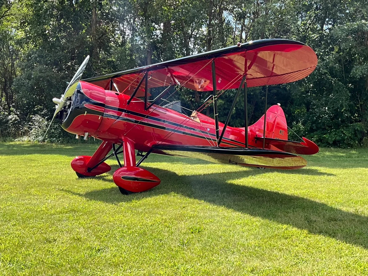 A red biplane with black accents and gold pinstripes is parked on a grassy field with trees in the background.