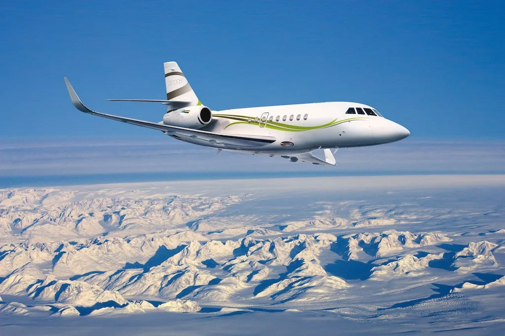 A white Dassault Falcon 2000S business jet with green and grey livery flies over snow-covered mountains under a clear blue sky.