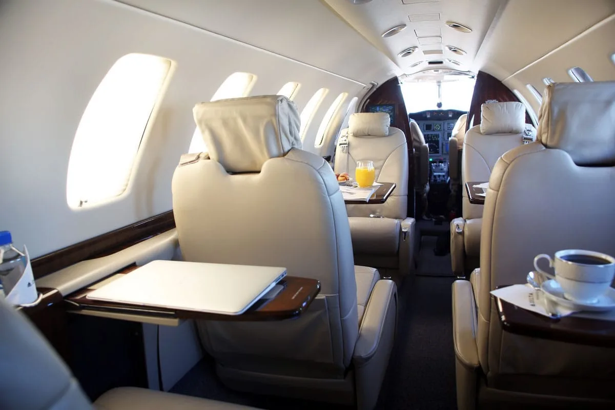 A view down the aisle of a private jet's cabin, showing beige leather seats, fold-out tables with a laptop and a cup of coffee.