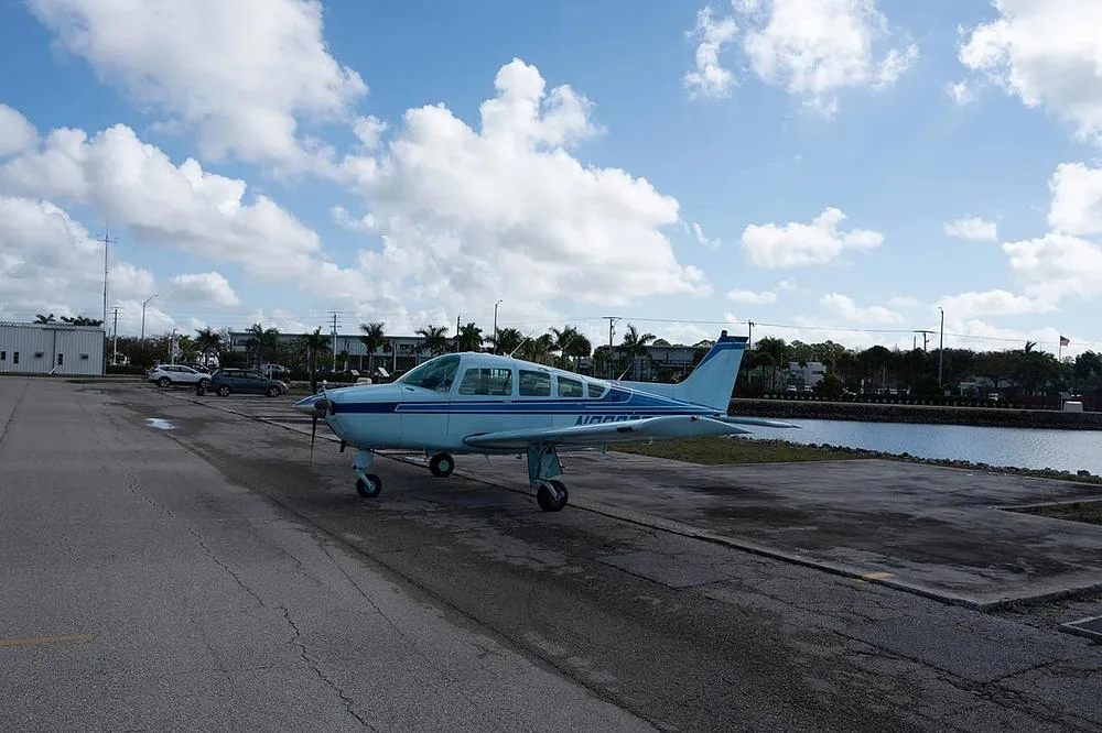 A light blue single-engine propeller aircraft, N9207S, is parked on an asphalt apron next to a body of water under a partly cloudy sky.