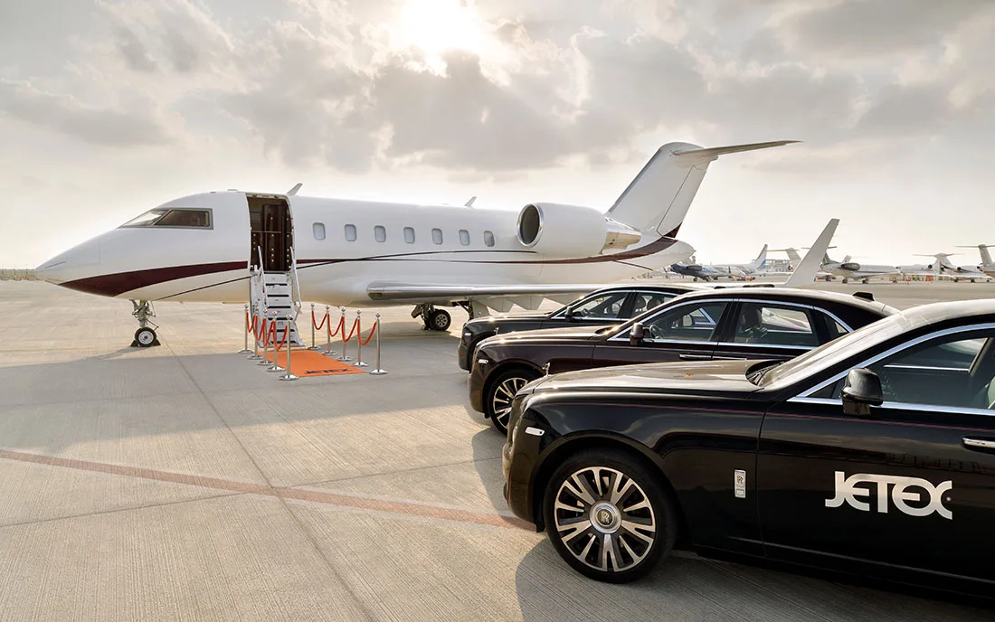 A white business jet with its door open and steps down, parked on a tarmac next to two black luxury cars, one with 'Jetex' branding.