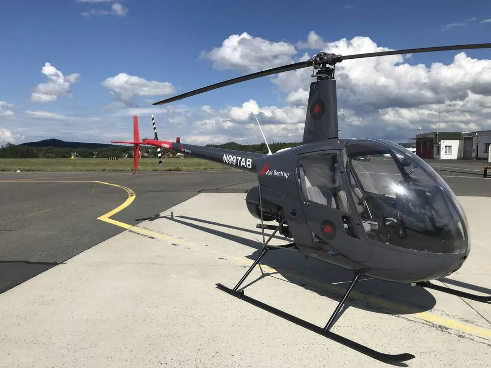 A dark gray Robinson R44 helicopter, N997AB, with red tail accents, parked on an airfield under a partly cloudy sky.