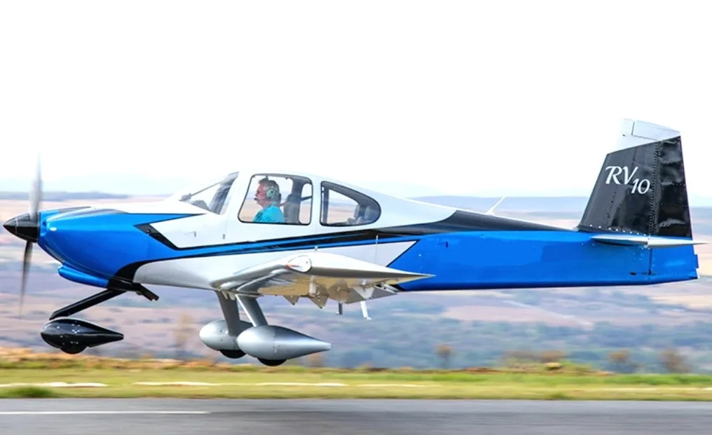 A blue, silver, and black Van's RV-10 aircraft with a pilot, in flight over a landscape.