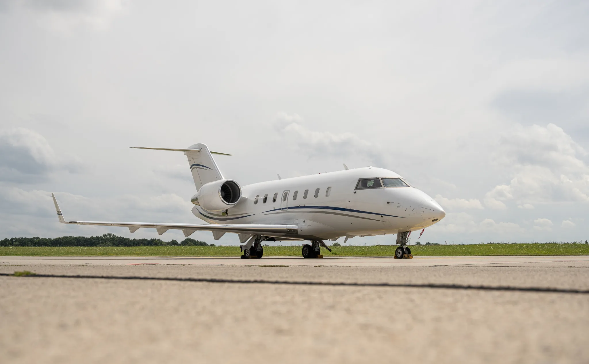 A white Bombardier Challenger business jet with blue and grey stripes on the tarmac under a cloudy sky.