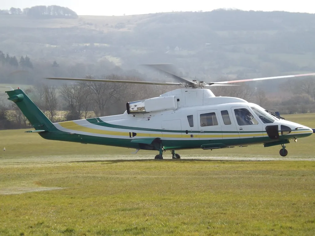 A white helicopter with green and yellow stripes is seen on a grassy field with hills in the background.
