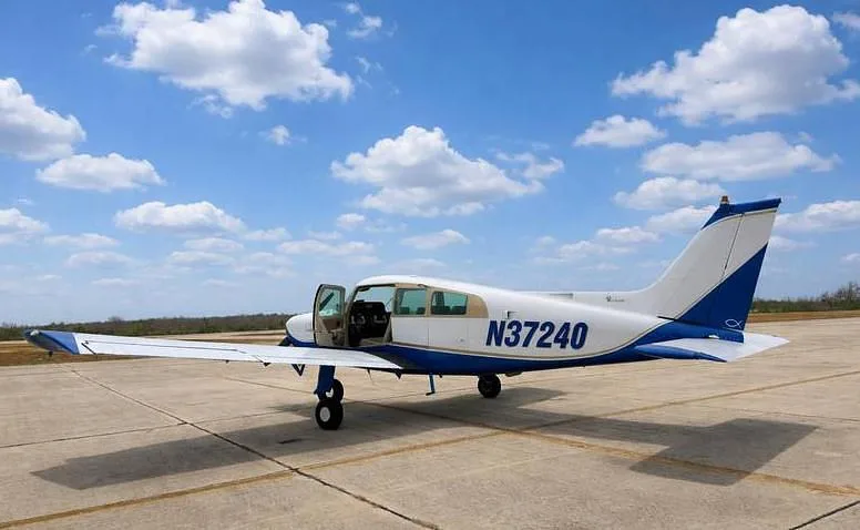 A side view of a white and blue Beechcraft Sundowner N37240 with its door open, parked on a tarmac.