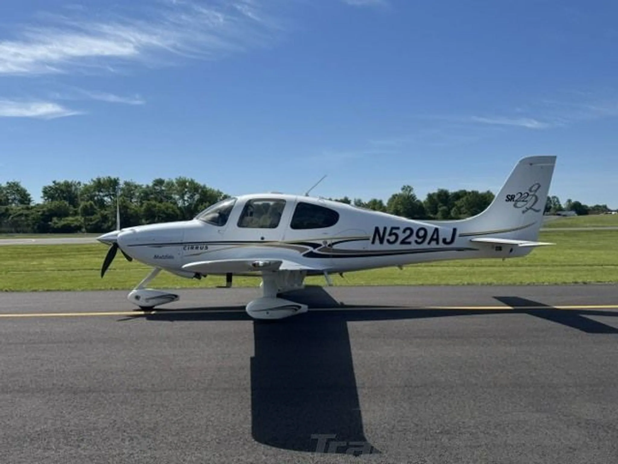 A white Cirrus SR22 aircraft with registration N529AJ parked on an asphalt runway under a bright blue sky.